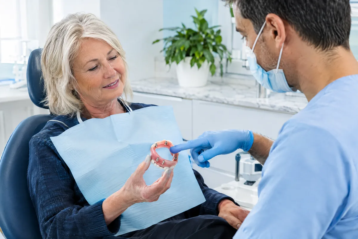 Older adult patient holding a removable denture while a dentist explains how it clips onto implants in a bright office